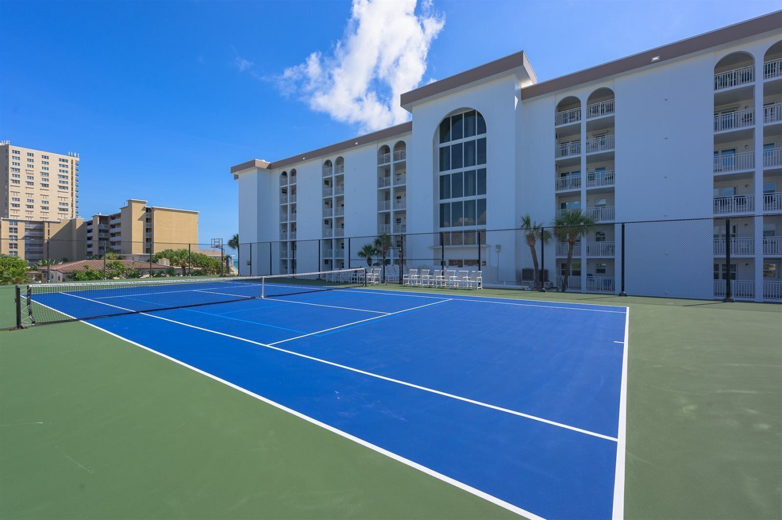 An outdoor tennis court with a net, surrounded by a fence, and adjacent to a building with multiple balconies.