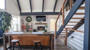 A spacious kitchen with a wooden island, two stools, a TV mounted on the wall, and a staircase leading to an upper level.