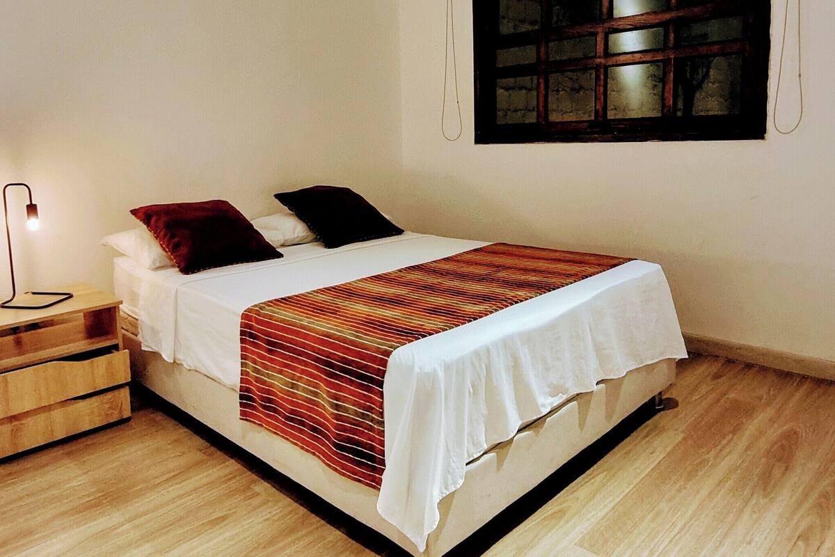 A bedroom with a bed, a bedside table, a lamp, and a window.