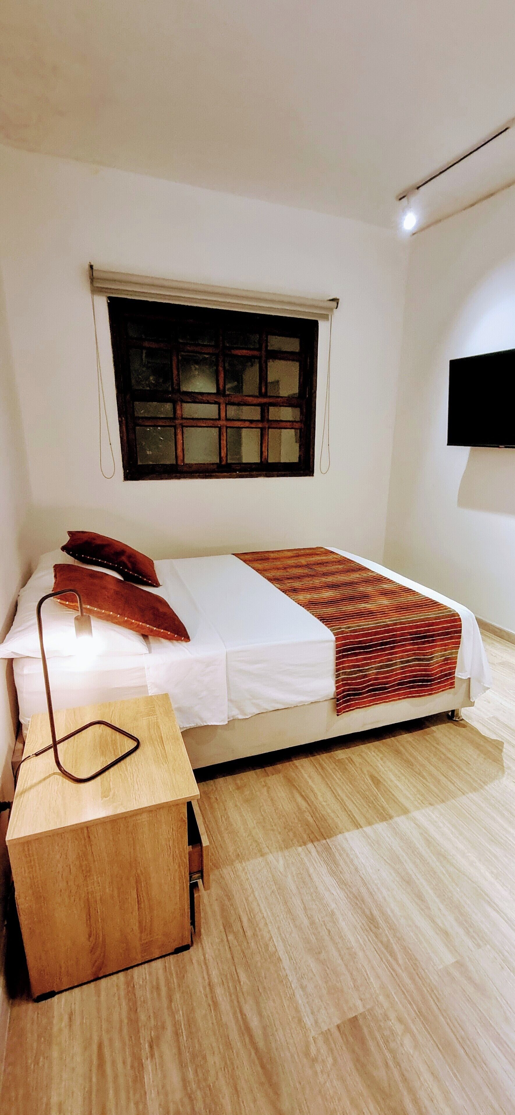 Una cama individual con colchón blanco y una manta estampada, una mesita de noche de madera con una lámpara, una ventana con cortina y un televisor de pantalla plana montado en la pared.