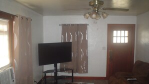 Een woonkamer met een flatscreen televisie op een standaard, een plafondventilator, een houten salontafel, een bruine bank en een raam met gordijnen.