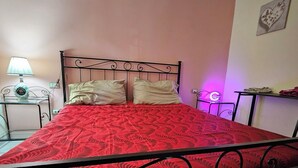 En seng med et rødt mønstret sengetæppe, en metalgavl og to puder. Der er en lampe på et natbord til venstre og et lille bord med en lampe til højre.