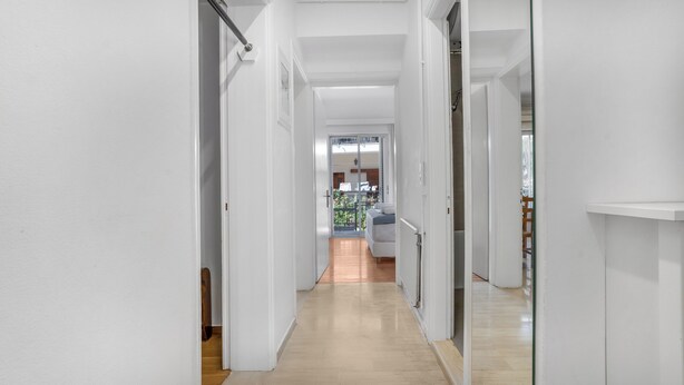Een smalle gang met witte wanden en houten vloeren die uitkomt op een balkon met uitzicht op groen.