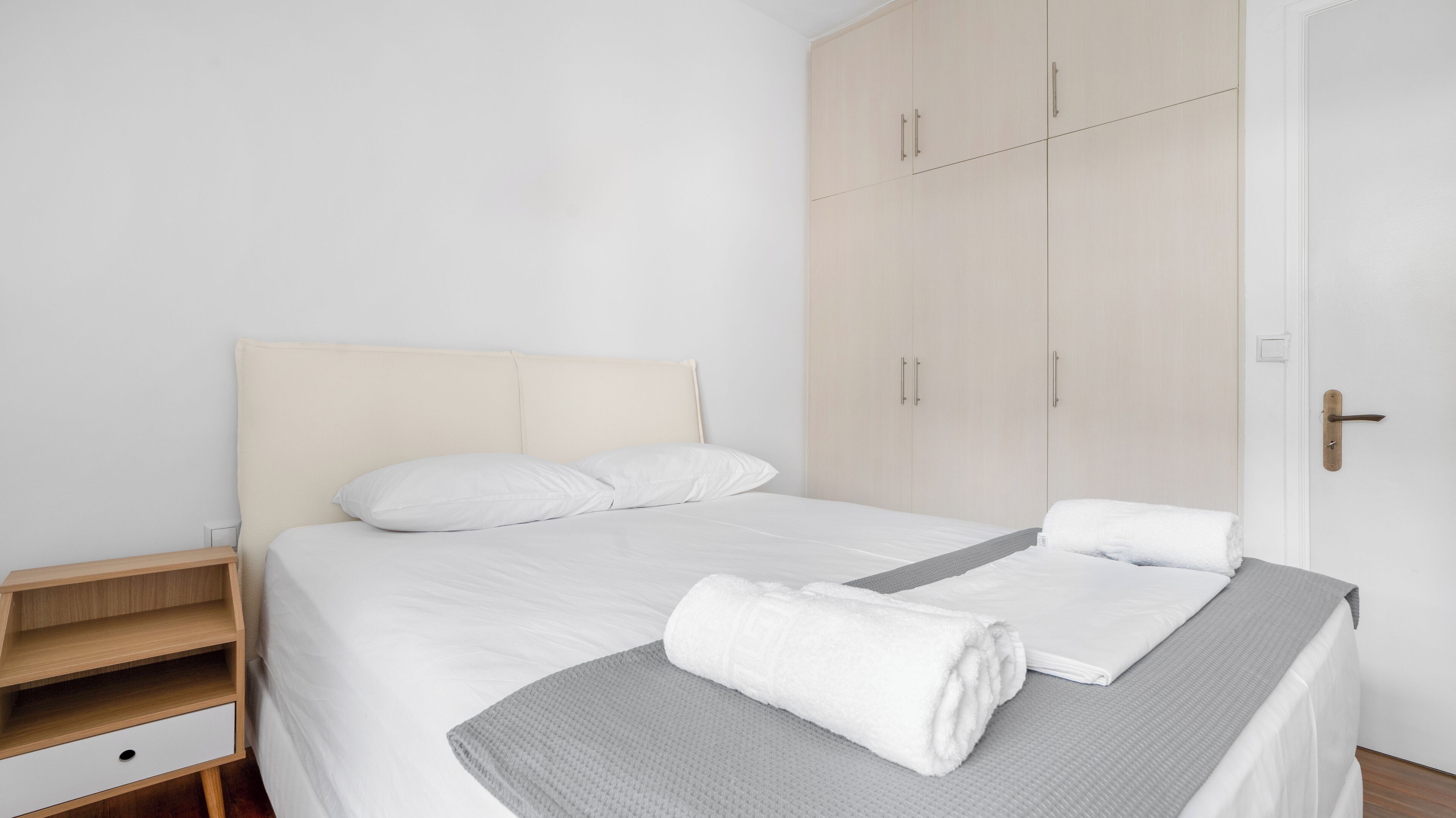 Ein ordentlich bezogenes Bett mit weißen Leinen, einer grauen Decke und einem aufgerollten weißen Handtuch. Ein Holznachttisch mit Schublade und kleiner Ablagefläche ist zu sehen. Im Hintergrund ist ein großer Kleiderschrank mit zwei Türen und Griffen erkennbar.
