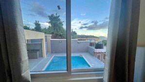 A view of a swimming pool through a window, with a clear sky and distant trees. - Hibiscus Charm - 2BR-2BA w/Pool (Kingston)