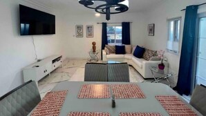 A modern living room with a flat-screen TV, a sofa set, a dining table with placemats, and a ceiling light fixture. - Hibiscus Charm - 2BR-2BA w/Pool (Kingston)