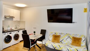 A compact living space with a kitchenette, a sofa with patterned cushions, a dining table, and a TV mounted on the wall. - Studio Jasmin (Paris)