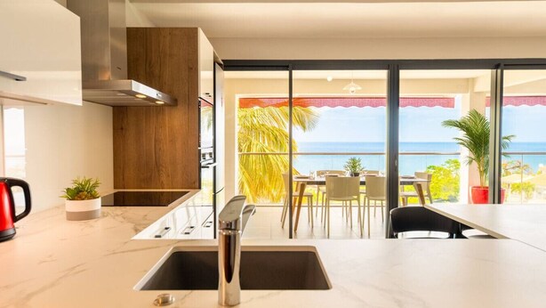 A modern kitchen with a large window overlooking the sea, a dining area with a table and chairs, and a view of the ocean. - Florida Sun : Beautiful appartment with sea view (Schoelcher)