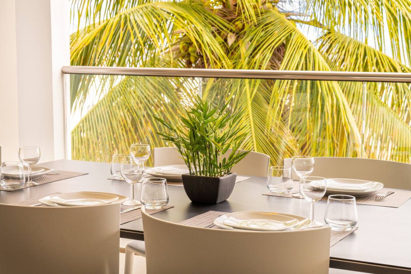 Mesa para comer con platos, vasos y una planta en maceta, con vista a palmeras a través de la barandilla de un balcón.