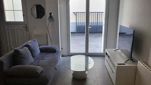 A modern living room with a sofa, a glass coffee table, a TV, and a sliding glass door leading to a balcony. - Appartement Proche de Vernon Giverny (Vernon)
