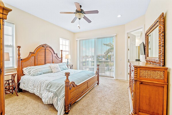 A bedroom with a wooden four-poster bed, a ceiling fan, a large window with blinds, a wooden dresser, and a mirror. - Beach Breezes & Backyard Bliss — Hot Tub, Fire Pit, Shuffleboard & BBQ Fun! 🏖️ (Indian Harbour Beach)