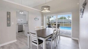 A dining area with a wooden table and white chairs, a kitchen in the background, and a view of a swimming pool through large glass doors. - Tropical Vacation Home Marco Island Florida - Close to Beaches-Pet Friendly-POOL (Marco Island)