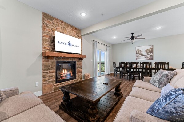 A living room with a stone fireplace, a dining area with a table and chairs, and a ceiling fan.