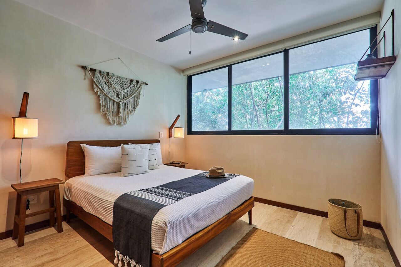 A bedroom with a bed, bedside lamps, a ceiling fan, a macrame wall hanging, and a large window with a view of trees.