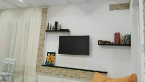 Un salon doté d’un téléviseur à écran plat fixé au mur, d’un canapé avec un coussin, d’un fauteuil et d’une étagère décorative où sont exposés divers objets.