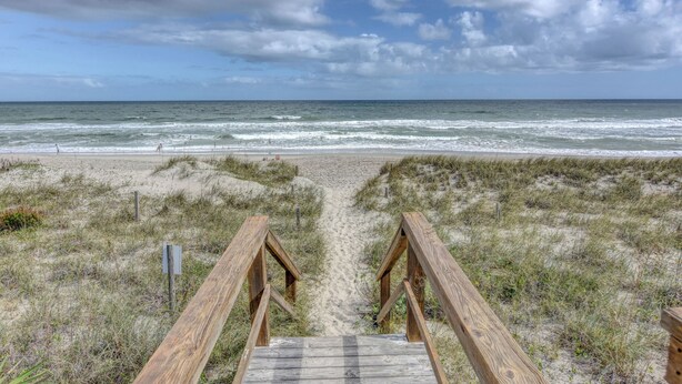 Een houten looppad dat naar een strand leidt, met een zandpad en grasduinen.