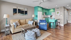 Een moderne woonkamer met een bank, salontafel, en poefs, een blauwe fauteuil, en een keuken met turquoise kastjes en een witte koelkast.