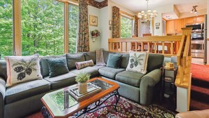 A cozy living room with a sectional sofa, a glass coffee table, and a view of lush greenery through large windows.