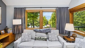 Un salon moderne avec un canapé gris, deux coussins décoratifs à motifs, une table d’appoint en bois avec une lampe, et une vue sur les arbres et les montagnes par une grande fenêtre.