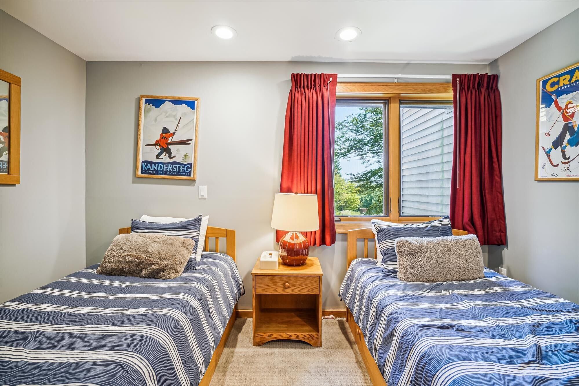 Un dormitorio con dos camas, una mesita de noche, una lámpara y una ventana con cortinas rojas.