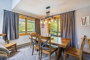 Un comedor con una mesa y sillas de madera, ventanas amplias con cortinas y una lámpara en el techo.