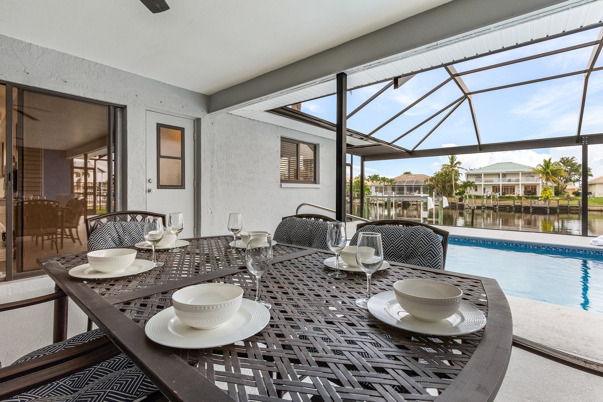A dining area with a pool view, featuring a table set with bowls and glasses, and a chair with a patterned cushion.