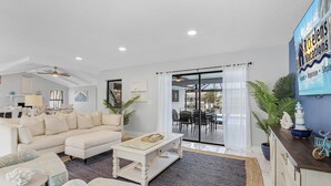 Un salon moderne avec un canapé, une table basse et un coin repas. On y trouve une grande fenêtre avec des rideaux, un ventilateur de plafond et une enseigne indiquant « Roelens Vacations ».