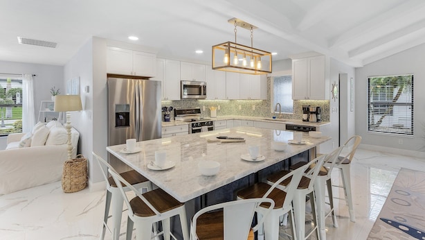Une cuisine moderne dotée d’un grand îlot central, de meubles blancs, d’appareils électroménagers en acier inoxydable et d’un luminaire suspendu.