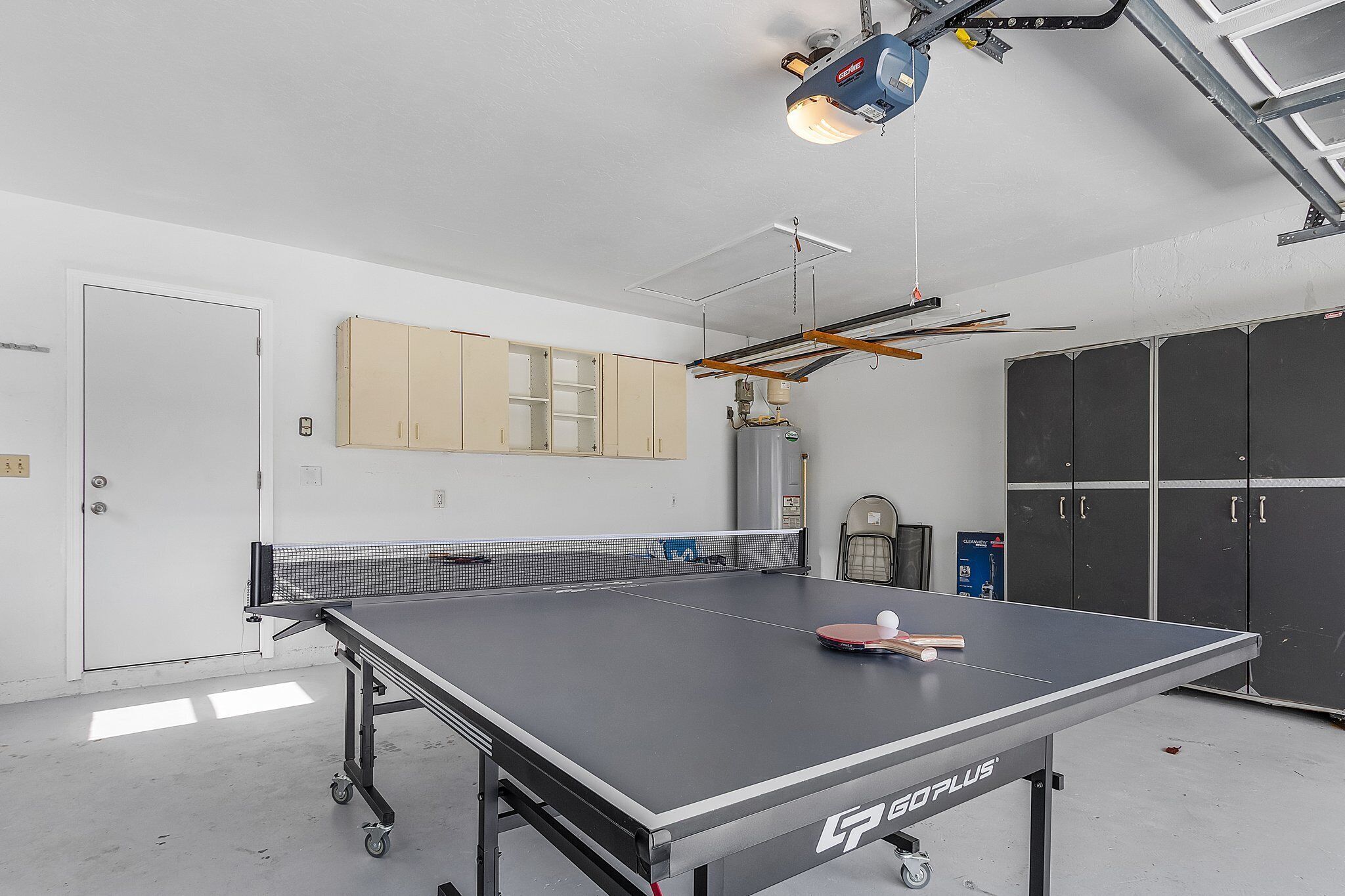 Una mesa de ping pong con red, una pelota y paletas en una habitación con paredes blancas, una puerta y armarios.