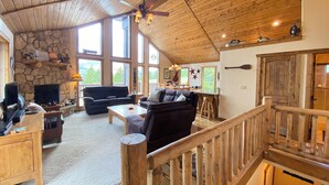 A cozy living room with a stone fireplace, wooden ceiling, and large windows offering a view of the outdoors. - Floor-to-Ceiling Mtn Views & Starlink - Elevated Deck, Fire Pit, Pet-Friendly (Fairplay)