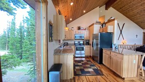 A modern kitchen with wooden cabinets, stainless steel appliances, and a large window overlooking a forest. - Floor-to-Ceiling Mtn Views & Starlink - Elevated Deck, Fire Pit, Pet-Friendly (Fairplay)