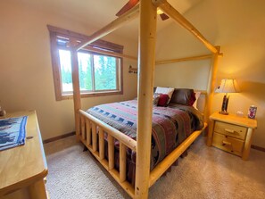Uma cama de dossel com colcha estampada, uma mesinha de cabeceira de madeira com abajur e uma janela com vista para árvores.