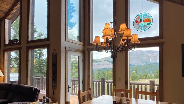 A spacious room with large windows offering mountain views, a wooden dining table with chairs, and a chandelier. - Floor-to-Ceiling Mtn Views & Starlink - Elevated Deck, Fire Pit, Pet-Friendly (Fairplay)