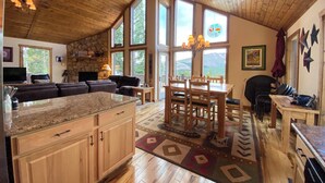 A spacious living area with a stone fireplace, wooden ceiling, and large windows offering a view of the outdoors. - Floor-to-Ceiling Mtn Views & Starlink - Elevated Deck, Fire Pit, Pet-Friendly (Fairplay)