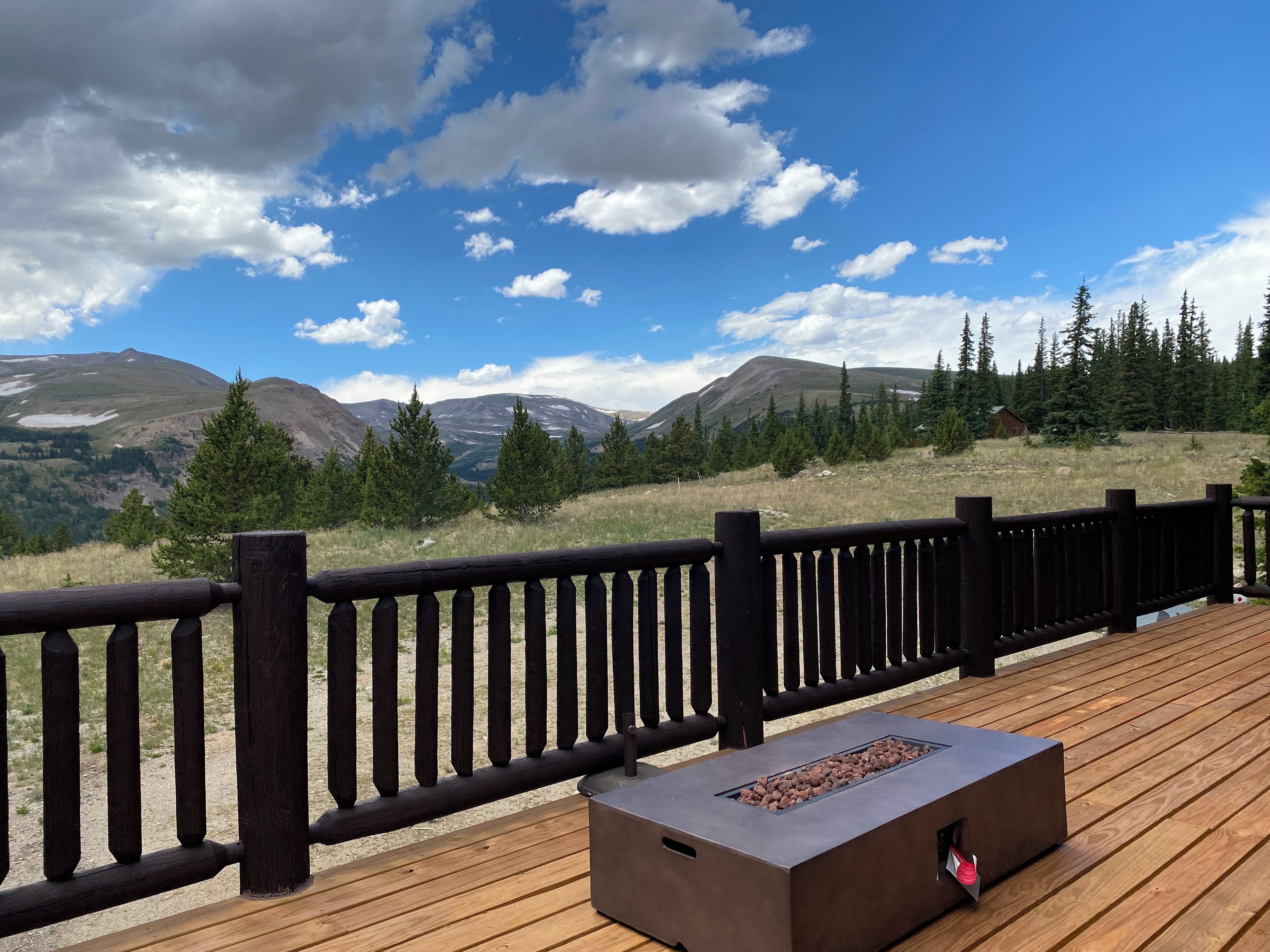 A wooden deck with a metal fire feature, overlooking a mountainous landscape with trees and a cabin.