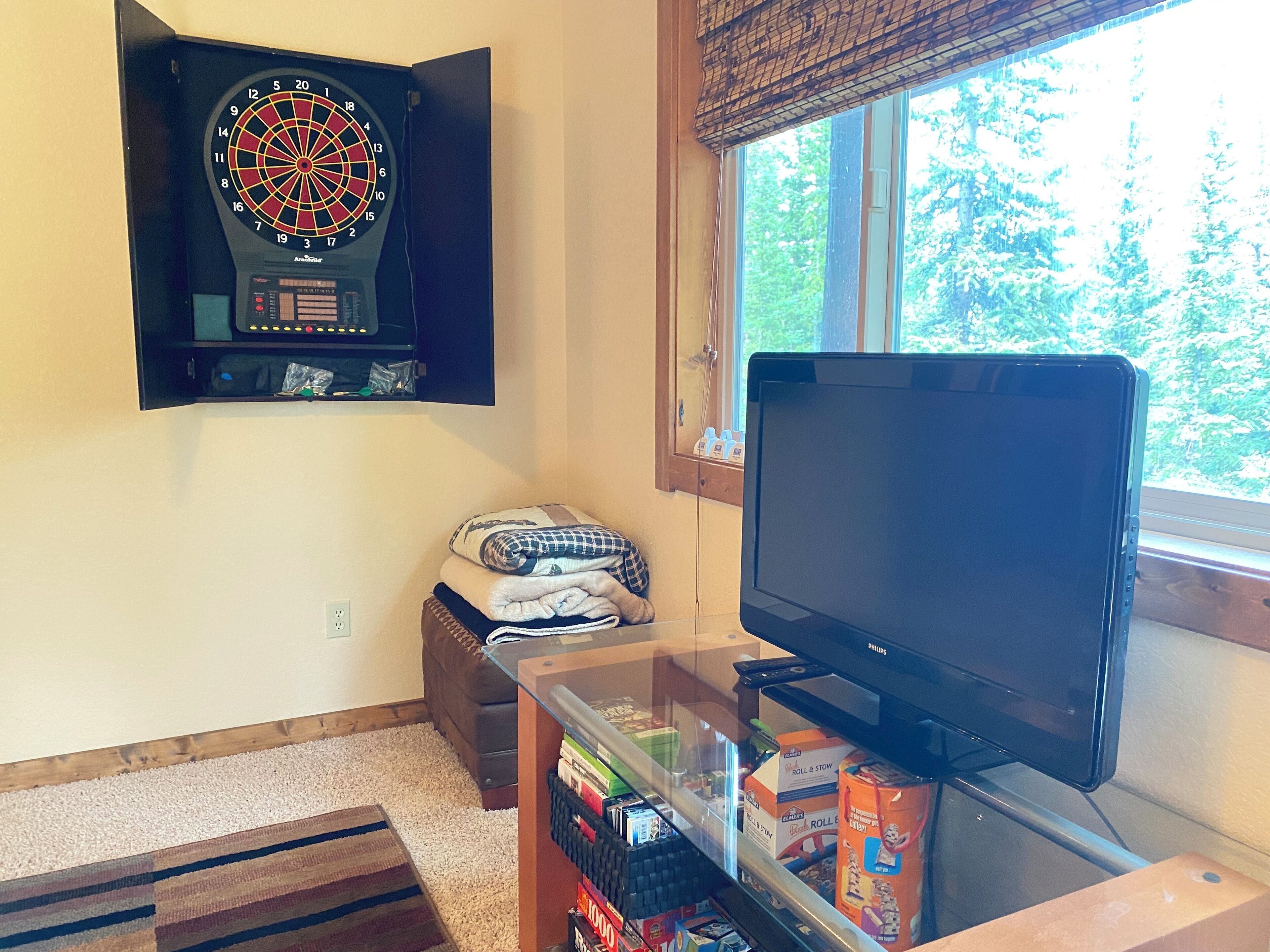 A room with a dartboard, a television, a window with a view of trees, and a shelf with books and a box.