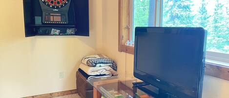 A room with a dartboard, a television, a window with a view of trees, and a shelf with books and a box.