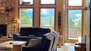 A cozy living room with a stone fireplace, leather sofa, and large windows offering mountain views. - Floor-to-Ceiling Mtn Views & Starlink - Elevated Deck, Fire Pit, Pet-Friendly (Fairplay)