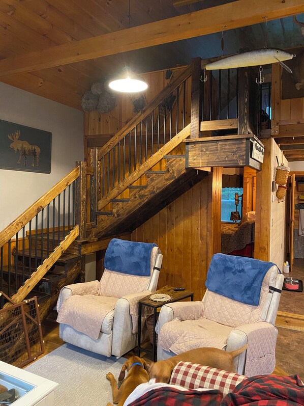 A cozy cabin interior with a wooden staircase, two cushioned chairs, a dog, and a bed with a checkered blanket.