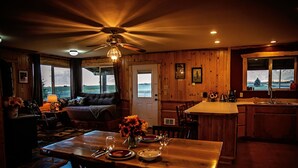 L’intérieur chaleureux d’un chalet comprend un coin repas, une cuisine et un salon, avec des murs en bois et de grandes fenêtres donnant sur un paysage magnifique.