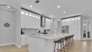 A modern kitchen with white cabinetry, a large island with bar stools, and a stainless steel refrigerator. - Ravello by the Beach - Beautiful pool home! (Naples)