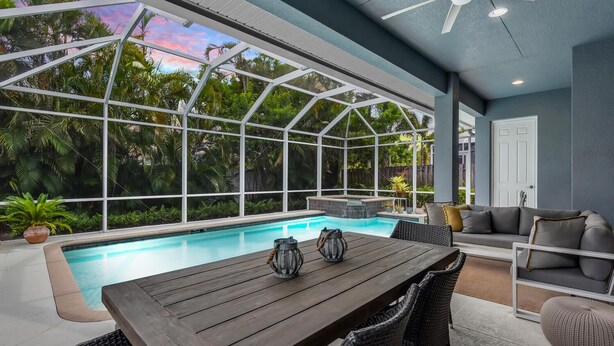 A covered outdoor pool area with a wooden table and chairs, a sofa, and a ceiling fan. - Ravello by the Beach - Beautiful pool home! (Naples)