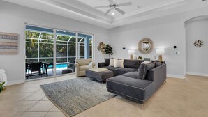 A spacious living room with a grey sectional sofa, a beige armchair, and a coffee table. There is a large window providing a view of a swimming pool and greenery outside. - Ravello by the Beach - Beautiful pool home! (Naples)