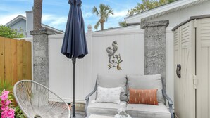 Eine Terrasse mit einem weißen Korbstuhl, einem grauen Sofa mit Kissen, einem Tisch und einem blauen Sonnenschirm.