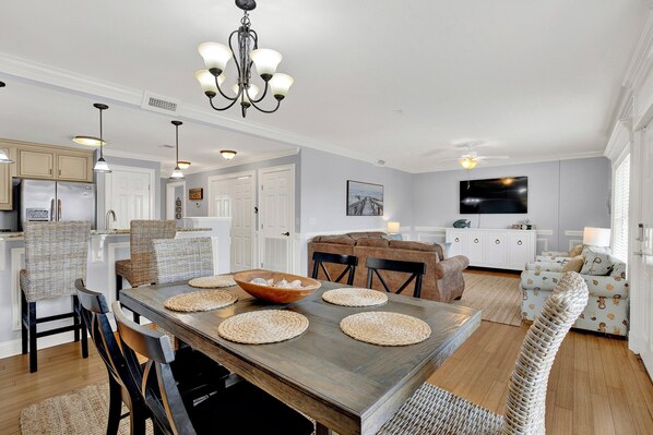 A modern kitchen with white cabinetry, a dining area with a wooden table, and a living room with a sofa and TV.
