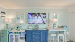 A room with a blue cabinet, a white desk, a TV, and a beach-themed painting. - Daytona Beach Studio w/ Ocean View + Amenities! (Daytona Beach)