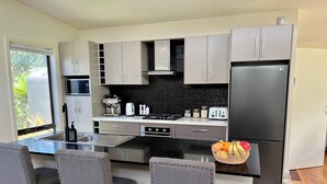A modern kitchen with grey cabinets, a black countertop, and a stainless steel refrigerator. There are three grey upholstered chairs with wooden legs and a black sink. - Banksia - EV charger and Linen provided (Venus Bay)