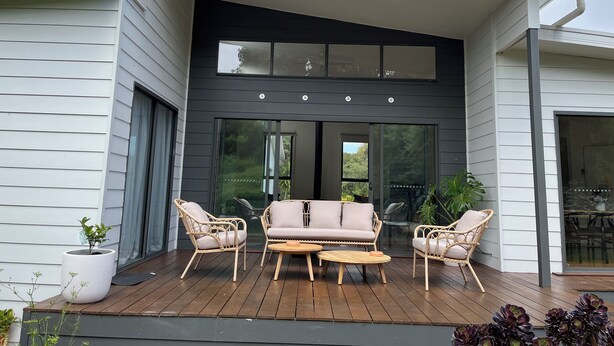 A modern house with a wooden deck, a black-framed glass door, and wicker furniture. - Banksia - EV charger and Linen provided (Venus Bay)