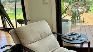 A modern living room with a beige cushioned chair, a small round table with a remote control, and a potted plant. - Banksia - EV charger and Linen provided (Venus Bay)