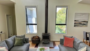 A living room with two grey sofas, a coffee table, a fireplace, and large windows with views of greenery. - Banksia - EV charger and Linen provided (Venus Bay)